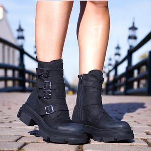 May include: Black ankle boots with multiple straps and buckles. The boots have a chunky sole and a side zipper. The boots are made of a matte material and are worn outdoors on a brick surface.