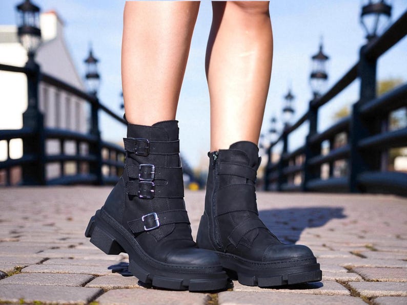 May include: Black ankle boots with multiple straps and buckles. The boots have a chunky sole and a side zipper. The boots are made of a matte material and are worn outdoors on a brick surface.