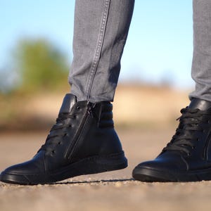 May include: Black leather high-top sneakers with a side zipper and black laces. The sneakers have a thick, textured sole and are worn with gray pants. The image is taken outdoors with a blurred background.