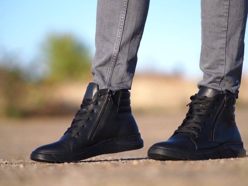 May include: Black leather high-top sneakers with a side zipper and black laces. The sneakers have a thick, textured sole and are worn with gray pants. The image is taken outdoors with a blurred background.