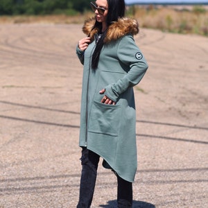 May include: A long, asymmetrical, hooded sweatshirt with a faux fur trim around the hood. The sweatshirt is a muted green color and has a zipper closure. The model is wearing black jeans and black sneakers.