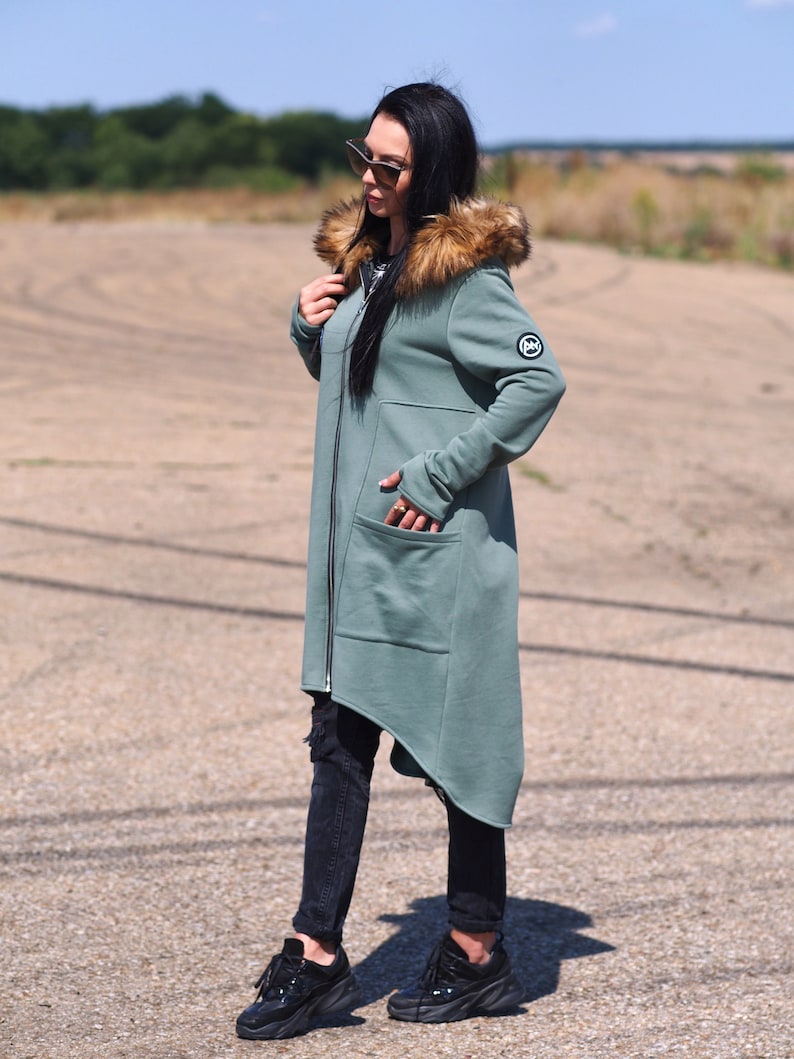 May include: A long, asymmetrical, hooded sweatshirt with a faux fur trim around the hood. The sweatshirt is a muted green color and has a zipper closure. The model is wearing black jeans and black sneakers.