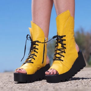 Könnte beinhalten: Ein Paar knöchelhohe Stiefel in leuchtendem Gelb mit offenem Zehen- und Fersenbereich. Die Stiefel verfügen über schwarze Schnürsenkel, einen seitlichen Reißverschluss und eine dicke, klobige schwarze Plateausohle mit tiefem Profil.