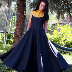 May include: A woman wearing a sleeveless dress with a yellow collar and a black skirt. The dress has a full skirt and is flowing in the wind. The woman is standing in a grassy area with trees in the background.