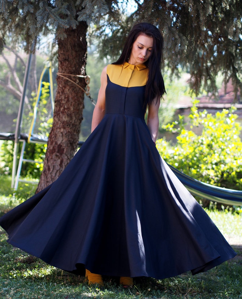 May include: A woman wearing a sleeveless dress with a yellow collar and a black skirt. The dress has a full skirt and is flowing in the wind. The woman is standing in a grassy area with trees in the background.