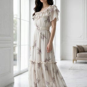 May include: A cream-colored maxi dress with a floral pattern. The dress features a square neckline, ruffled sleeves, and a belted waist. The skirt has a tiered design. The model is wearing silver heels.