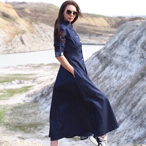 May include: A woman wearing a dark blue denim dress with a button-up front and long sleeves. She is wearing sunglasses and white sneakers with black accents. The woman is standing on a rocky surface with a body of water in the background.