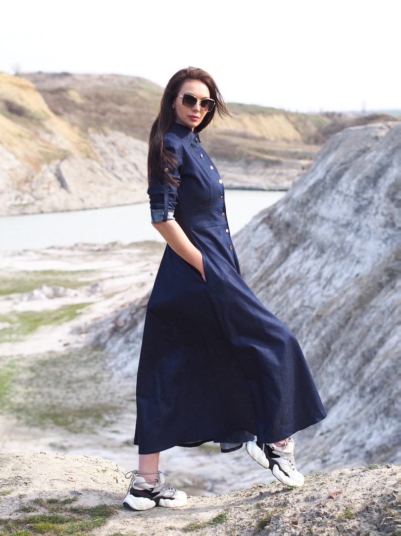 May include: A woman wearing a dark blue denim dress with a button-up front and long sleeves. She is wearing sunglasses and white sneakers with black accents. The woman is standing on a rocky surface with a body of water in the background.