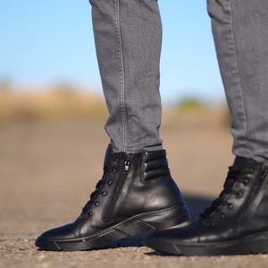 May include: Black leather ankle boots with a side zipper and lace-up closure. The boots have a ridged sole and are worn with gray denim jeans. The boots are a solid black color.