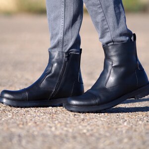 May include: Black leather ankle boots with a side zipper and a rounded toe. The boots have a low, dark sole and are worn with gray pants. The boots appear to be new and unworn.