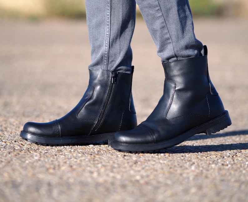 May include: Black leather ankle boots with a side zipper and a rounded toe. The boots have a low, dark sole and are worn with gray pants. The boots appear to be new and unworn.