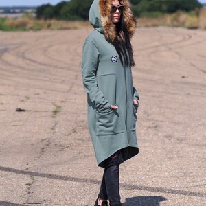 May include: A long, sage green hooded sweatshirt with a faux fur hood. The sweatshirt has a zipper closure and a small logo patch on the chest. The model is wearing black jeans and black sneakers.