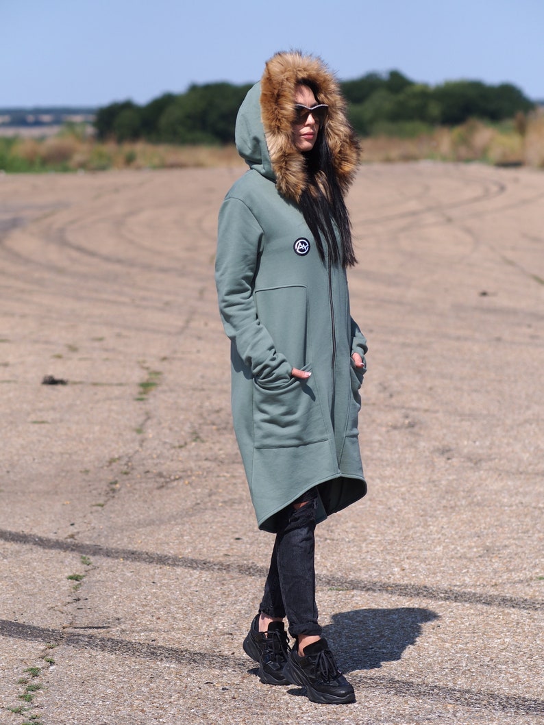 May include: A long, sage green hooded sweatshirt with a faux fur hood. The sweatshirt has a zipper closure and a small logo patch on the chest. The model is wearing black jeans and black sneakers.
