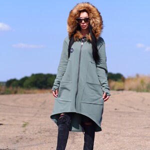 May include: A long, green hooded sweatshirt with a faux fur hood. The sweatshirt has a zipper closure and two large pockets. The model is wearing black jeans and black sneakers.
