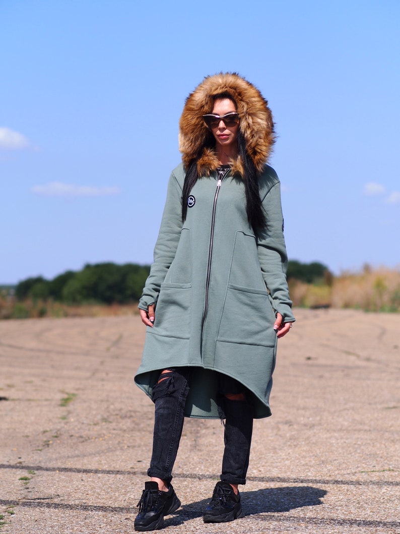 May include: A long, green hooded sweatshirt with a faux fur hood. The sweatshirt has a zipper closure and two large pockets. The model is wearing black jeans and black sneakers.