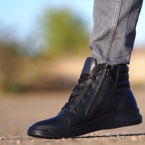 May include: Black high-top sneaker with a side zipper and laces. The shoe has a smooth leather upper and a black sole. The design includes a padded ankle collar and a textured heel panel. The sneaker is worn with gray jeans.