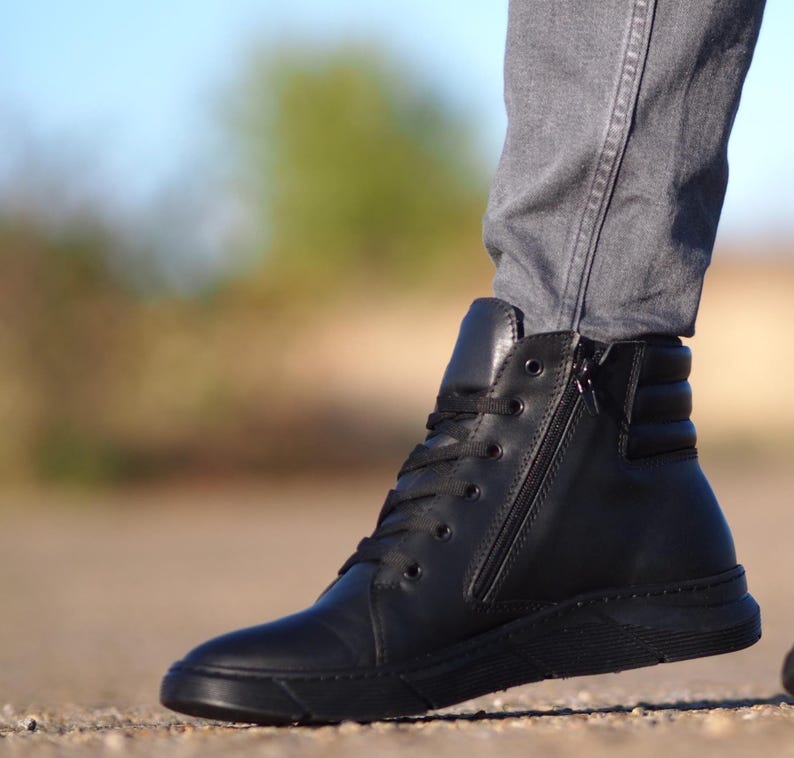 May include: Black high-top sneaker with a side zipper and laces. The shoe has a smooth leather upper and a black sole. The design includes a padded ankle collar and a textured heel panel. The sneaker is worn with gray jeans.