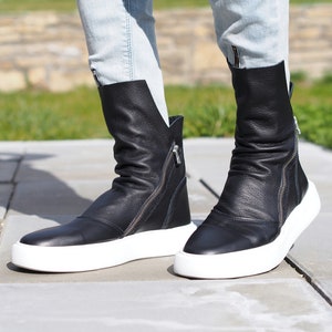 Puede incluir: Botas de tobillo de cuero negro con suelas blancas y cremalleras laterales. Las botas est&aacute;n ligeramente ca&iacute;das y tienen un aspecto informal y deportivo.