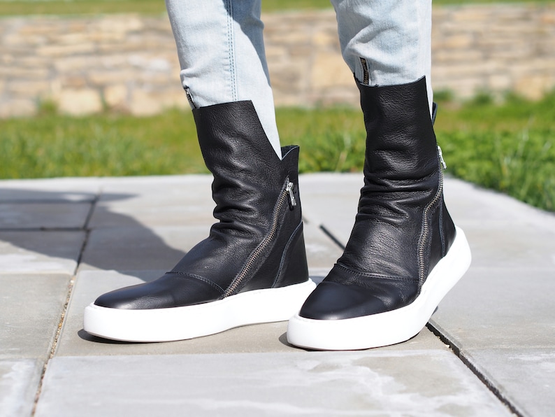 Puede incluir: Botas de tobillo de cuero negro con suelas blancas y cremalleras laterales. Las botas est&aacute;n ligeramente ca&iacute;das y tienen un aspecto informal y deportivo.