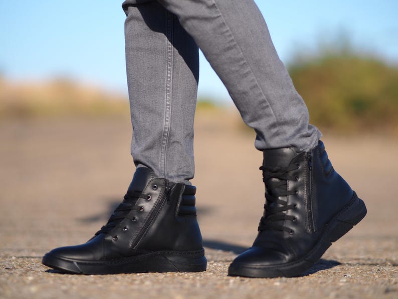 May include: Black leather high-top sneakers with side zippers and lace-up closures. The shoes have a thick, textured sole and are worn with gray jeans. The image is taken outdoors on a sunny day.