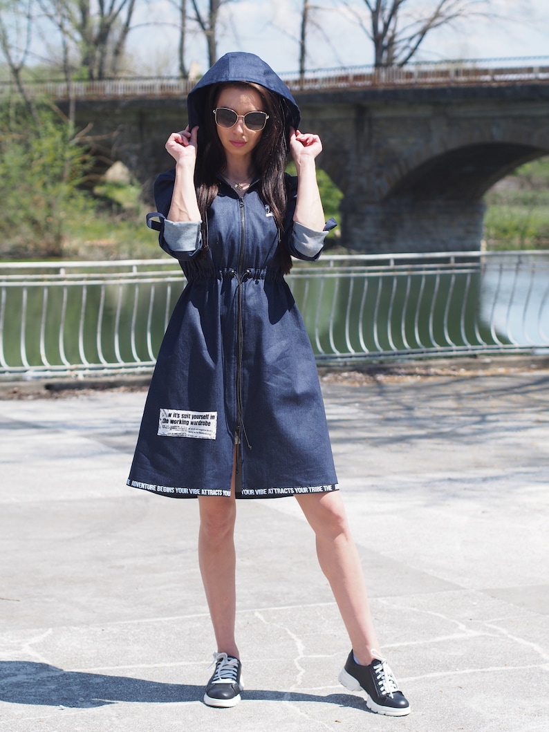 May include: A blue denim dress with a hood and a zipper closure. The dress has a drawstring waist and white text on the bottom hem that reads "Adventure Begins Your Vibe Attracts Your Tribe".