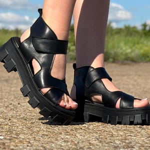Pu&ograve; includere: Sandali platform in pelle nera con suola carrarmato. I sandali hanno un design a punta aperta con pi&ugrave; cinturini e chiusura in velcro. Le scarpe sono indossate all'aperto su una superficie pavimentata.