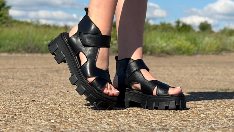 Pu&ograve; includere: Sandali platform in pelle nera con suola carrarmato. I sandali hanno un design a punta aperta con pi&ugrave; cinturini e chiusura in velcro. Le scarpe sono indossate all'aperto su una superficie pavimentata.