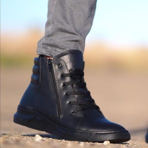 May include: Black high-top boot with a side zipper and black laces. The boot has a padded ankle and a thick, textured sole. The boot is made of a smooth, matte material and is worn with gray pants.