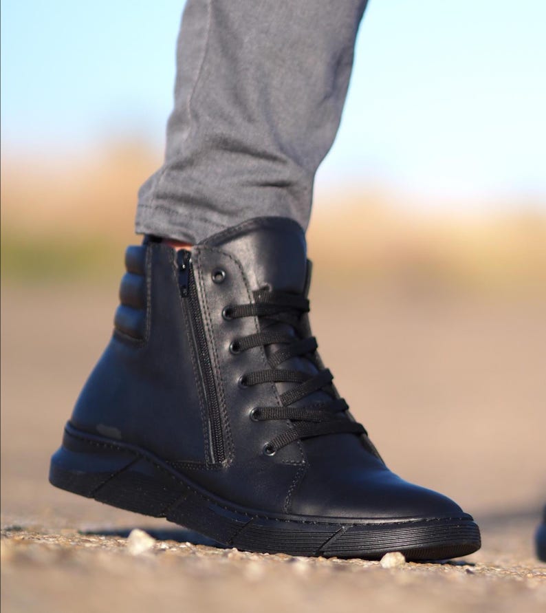 May include: Black high-top boot with a side zipper and black laces. The boot has a padded ankle and a thick, textured sole. The boot is made of a smooth, matte material and is worn with gray pants.