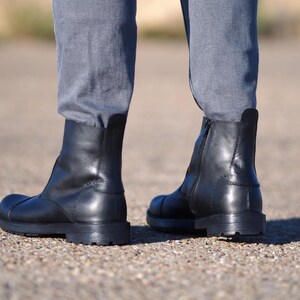 May include: A pair of black leather ankle boots with a rounded toe and a low, stacked heel. The boots have a zipper on the side and are worn with gray pants. The boots appear to be new and unworn.