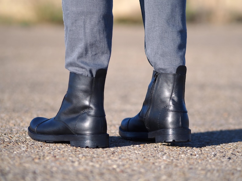 May include: A pair of black leather ankle boots with a rounded toe and a low, stacked heel. The boots have a zipper on the side and are worn with gray pants. The boots appear to be new and unworn.
