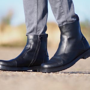 May include: Black leather boots with a side zipper and a rounded toe. The boots have a thick sole and are worn with gray pants. The boots appear to be new and unworn.