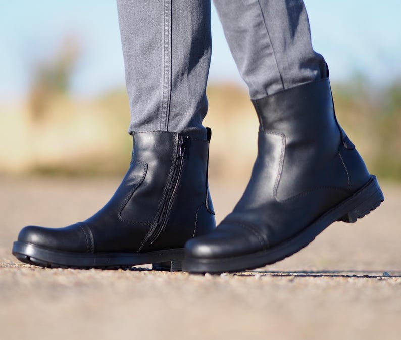 May include: Black leather boots with a side zipper and a rounded toe. The boots have a thick sole and are worn with gray pants. The boots appear to be new and unworn.
