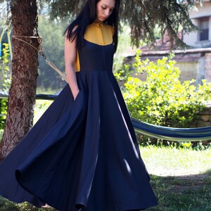May include: A woman wearing a navy blue maxi dress with a yellow collared shirt underneath. The dress has a full skirt and pockets on the sides. She is standing in a garden with a hammock in the background.