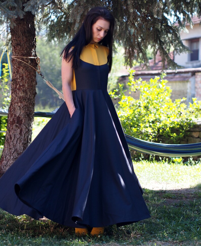 May include: A woman wearing a navy blue maxi dress with a yellow collared shirt underneath. The dress has a full skirt and pockets on the sides. She is standing in a garden with a hammock in the background.