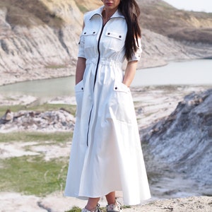 May include: A white maxi dress with a zipper down the front and a drawstring waist. The dress has short sleeves and a relaxed fit. The model is wearing the dress with white sneakers.