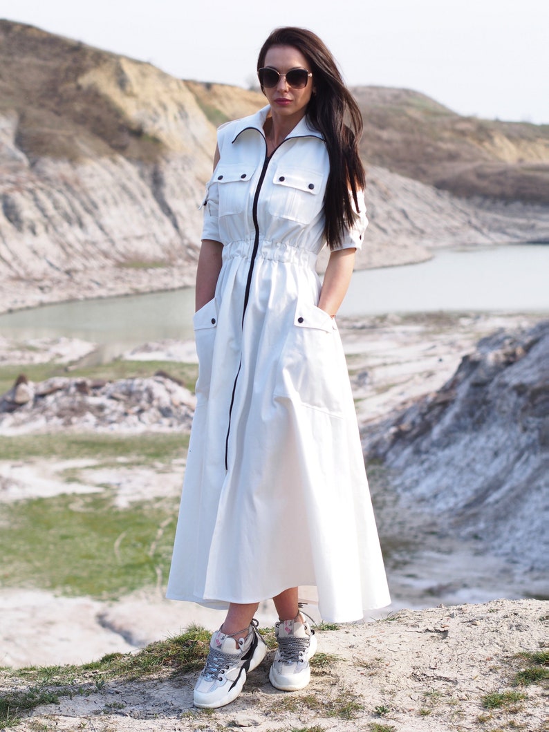 May include: A white maxi dress with a zipper down the front and a drawstring waist. The dress has short sleeves and a relaxed fit. The model is wearing the dress with white sneakers.