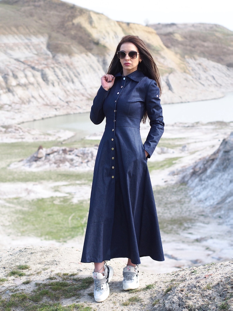 May include: A long, dark blue denim dress with a button-up front and a collared neckline. The dress has a flared skirt and is worn with white sneakers.