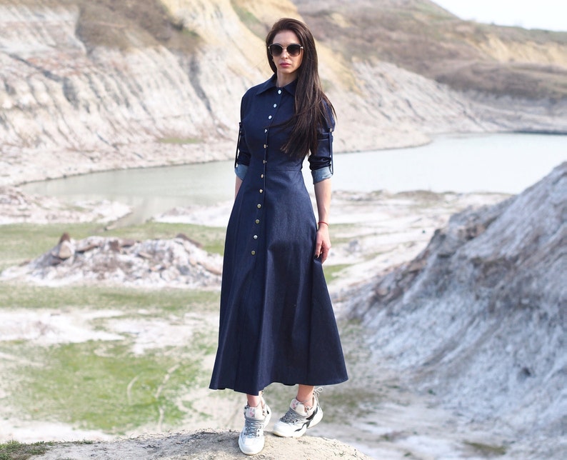 May include: A woman wearing a long, dark blue denim dress with a button-up front and long sleeves. She is wearing white sneakers and sunglasses.