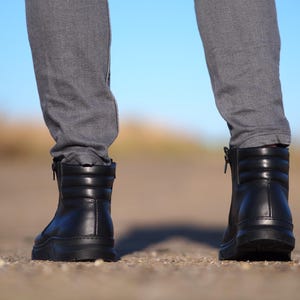 May include: Black leather boots with a quilted design and side zippers. The boots have a thick sole and are worn with gray pants. The background is a blurred view of a road and a blue sky.