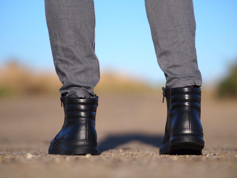 May include: Black leather boots with a quilted design and side zippers. The boots have a thick sole and are worn with gray pants. The background is a blurred view of a road and a blue sky.
