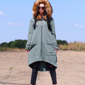 May include: A long, sage green hooded sweatshirt with a faux fur hood. The sweatshirt has a zipper closure and two large pockets. The model is wearing black jeans and black sneakers.