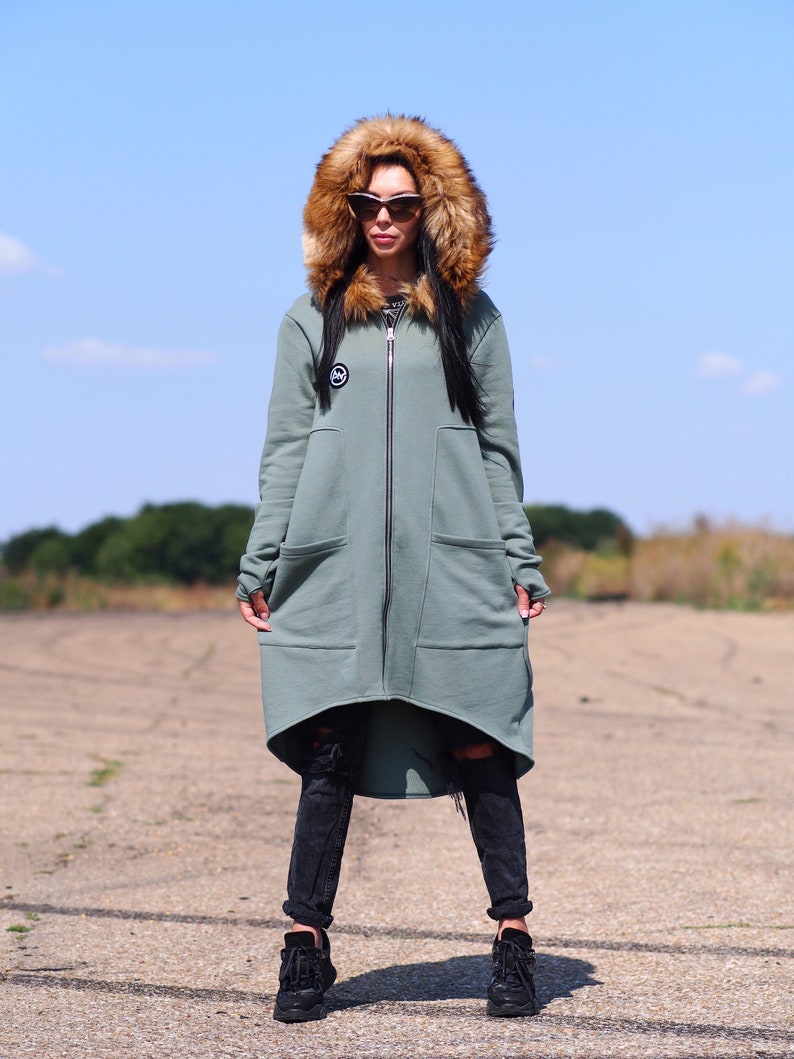 May include: A long, sage green hooded sweatshirt with a faux fur hood. The sweatshirt has a zipper closure and two large pockets. The model is wearing black jeans and black sneakers.