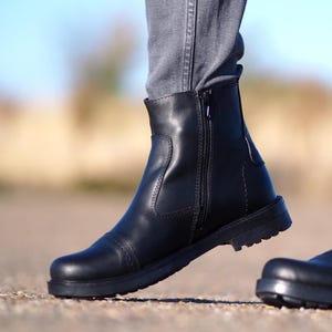 May include: Black leather ankle boot with a side zipper and a rounded toe. The boot has a low, thick sole and is worn with gray denim jeans. The boot's design is simple and classic, suitable for various outfits.
