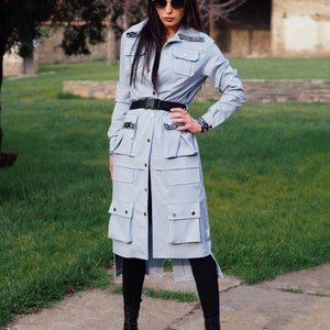 May include: A light blue long-sleeved shirt with a belt and multiple pockets. The shirt has a button-up front and a relaxed fit. The model is wearing black leggings and black shoes.