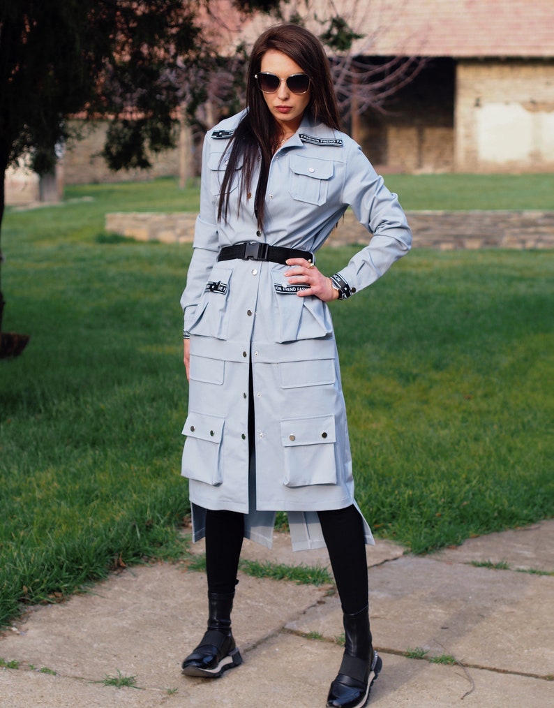 May include: A light blue long-sleeved shirt with a belt and multiple pockets. The shirt has a button-up front and a relaxed fit. The model is wearing black leggings and black shoes.