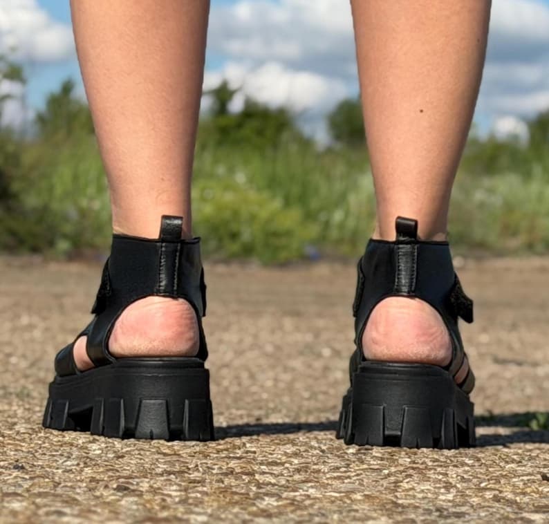 Pu&ograve; includere: Sandali neri con plateau e suola spessa. I sandali hanno il retro aperto e un cinturino alla caviglia. Le suole sono spesse circa 7,6 cm. I sandali sono indossati su una superficie di ghiaia.