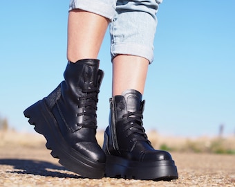May include: A pair of black leather ankle boots with chunky platform soles and lace-up fastenings. The boots have a side zip and are being worn by someone standing on a gravel path.