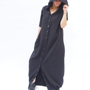 Puede incluir: Un vestido largo negro con capucha, con botones en la parte delantera y una falda larga y fluida. El vestido tiene un corte holgado y est&aacute; hecho de una tela suave y c&oacute;moda.