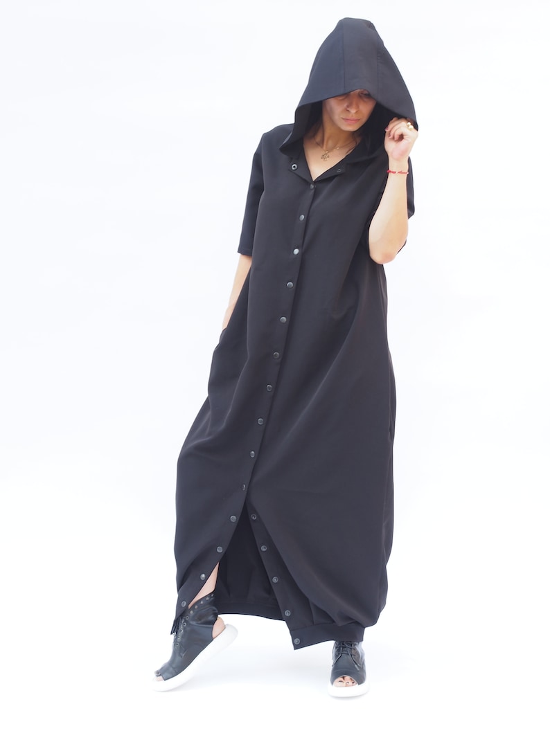 Puede incluir: Un vestido largo negro con capucha, con botones en la parte delantera y una falda larga y fluida. El vestido tiene un corte holgado y est&aacute; hecho de una tela suave y c&oacute;moda.